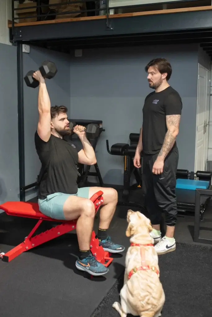 Adhérent réalisant un exercice supervisé par un coach pendant qu’Arya observe la séance